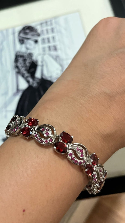Close-up of a hand wearing a sterling silver bracelet intricately set with oval-shaped red rubies and garnets, interspersed with silver swirl designs