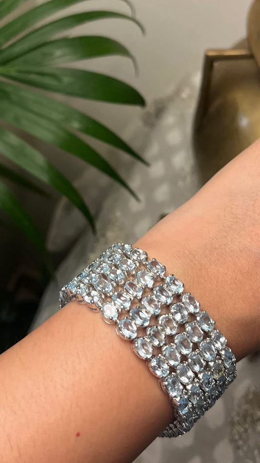 Close-up of a hand wearing a wide, multi-row silver bracelet densely set with many oval or round cut blue topaz gemstones, reflecting light.