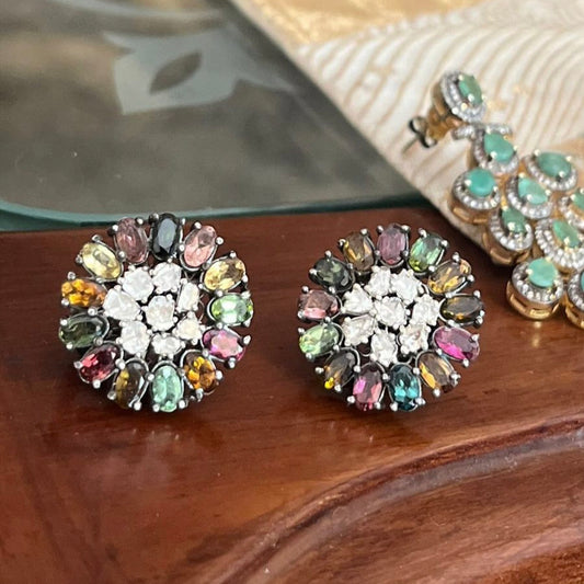 Close-up view of two round handmade stud earrings featuring a cluster of multi-colored tourmaline gemstones surrounding a central arrangement of polki diamonds, displayed on a wooden surface with a glimpse of another gold and emerald earring in the background.