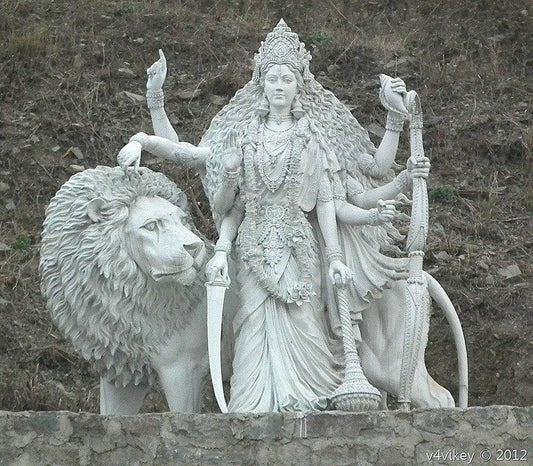 White marble statue of Goddess Durga with eight arms, holding various weapons, standing beside a majestic lion on a stone base, against a natural, rocky background.
